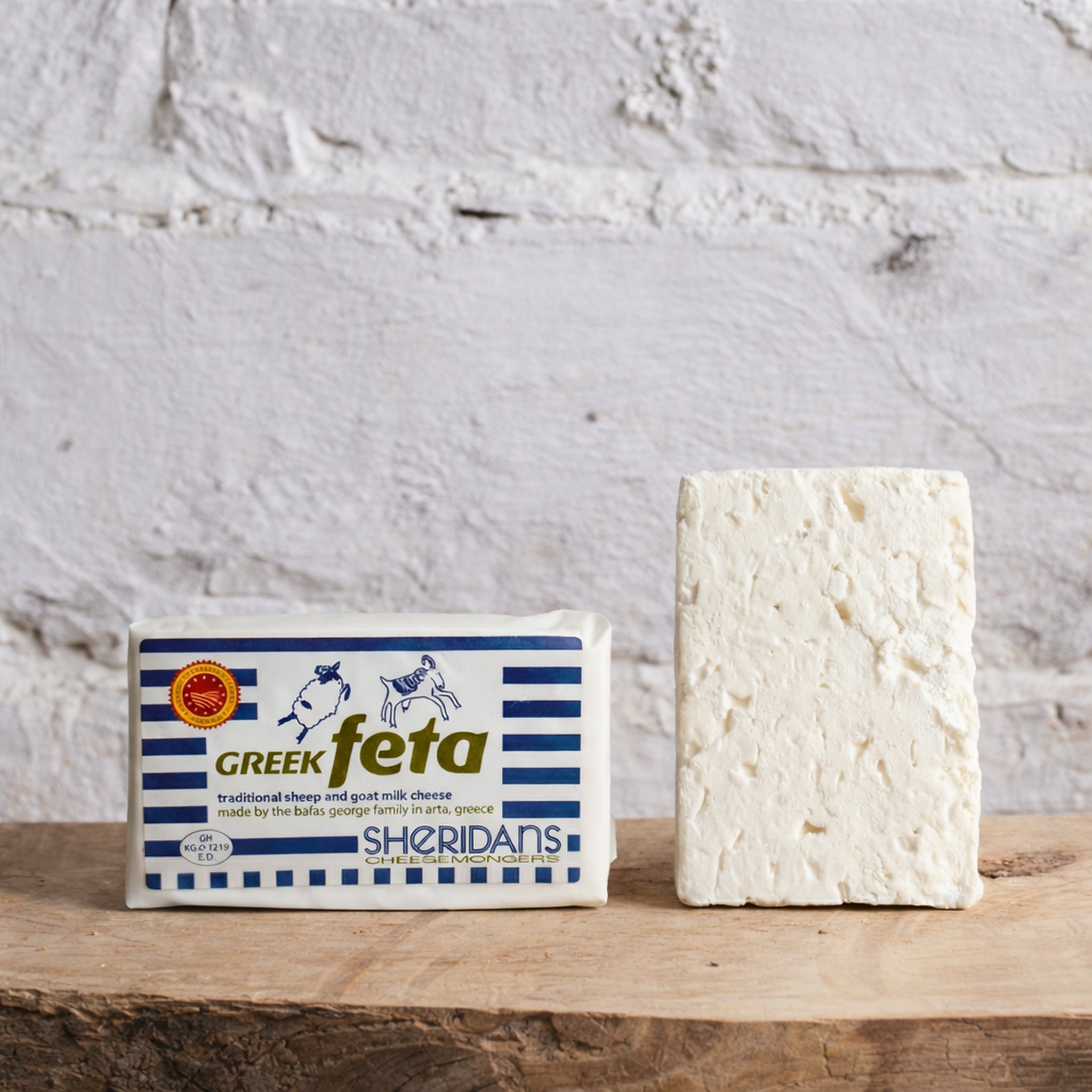 Block of Greek feta cheese next to its packaging on a wooden surface with a white brick wall background.