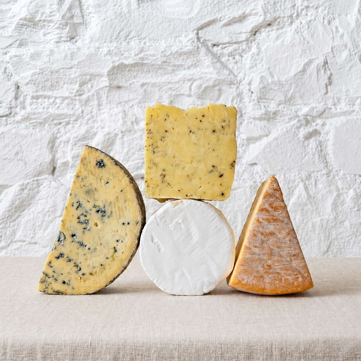 Four types of artisanal cheese displayed on a beige linen surface against a white textured brick wall. The assortment includes a wedge of blue cheese, a square slice of yellow cheese with herbs, a round wheel of soft white cheese, and a wedge of orange-brown hard cheese.