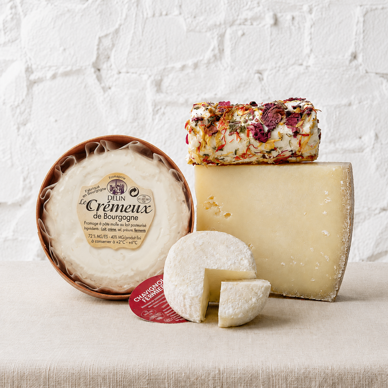 Assorted cheeses including a wheel of delice8 Le Creèmeux de Bourgogne, a block of Gorgonzola, and a wedge of Parmesan on a textured surface with a white brick wall background.