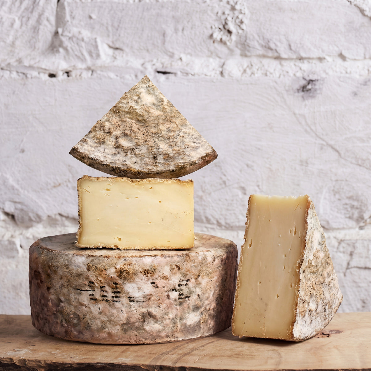 Three wedges and a block of artisanal cheese displayed on a wooden board against a white brick wall background.