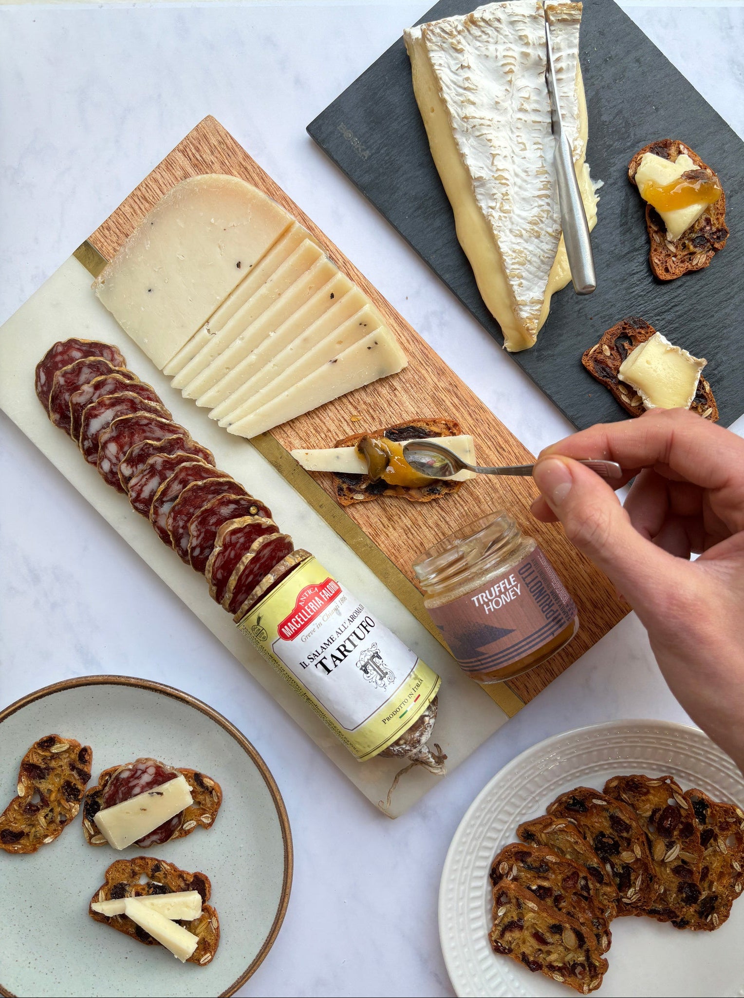 Platter of assorted cheeses, cured meats, and bread with a hand holding a jar of jam.