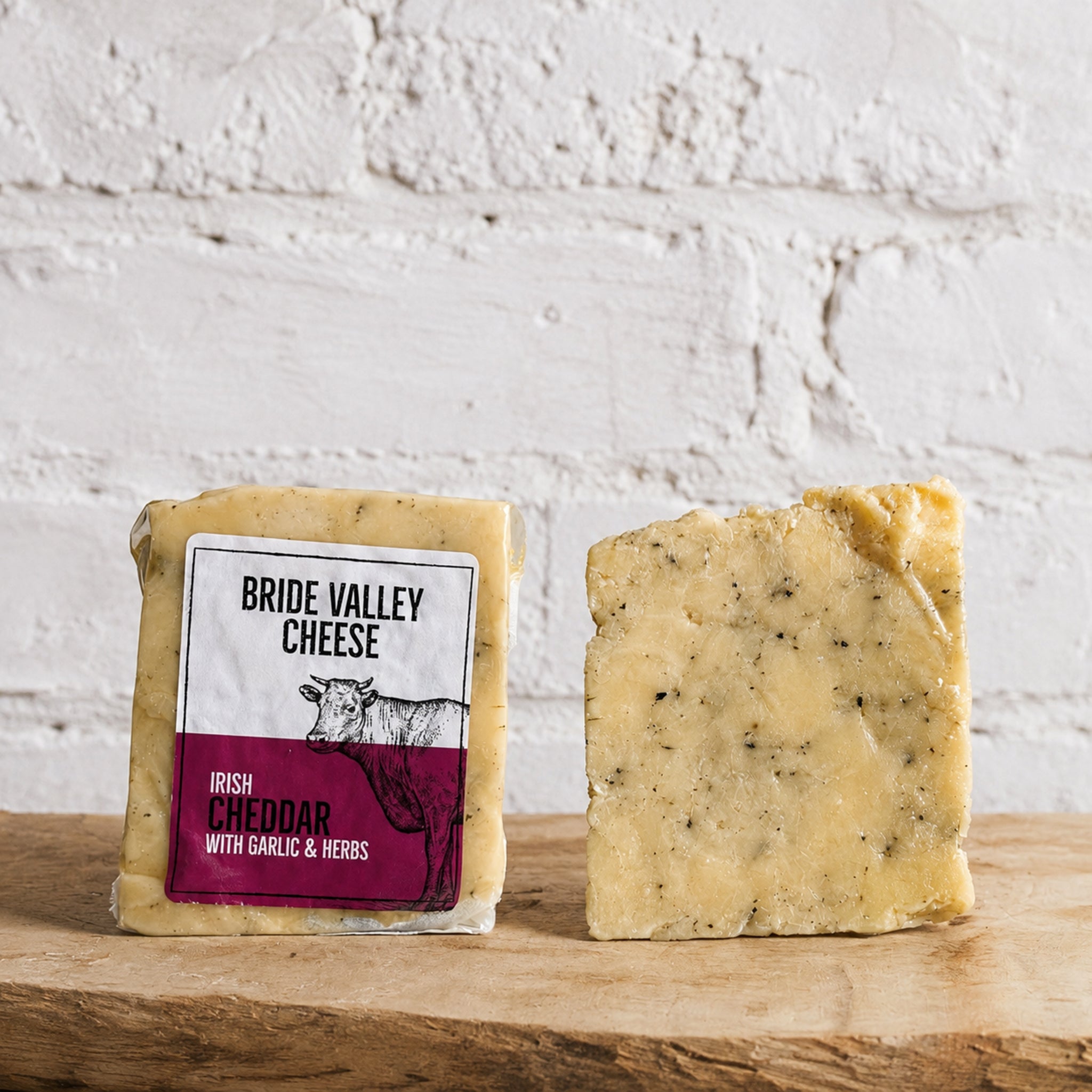 Bride Valley Cheese Irish Cheddar with Garlic & Herbs on a wooden surface with a white brick wall background.