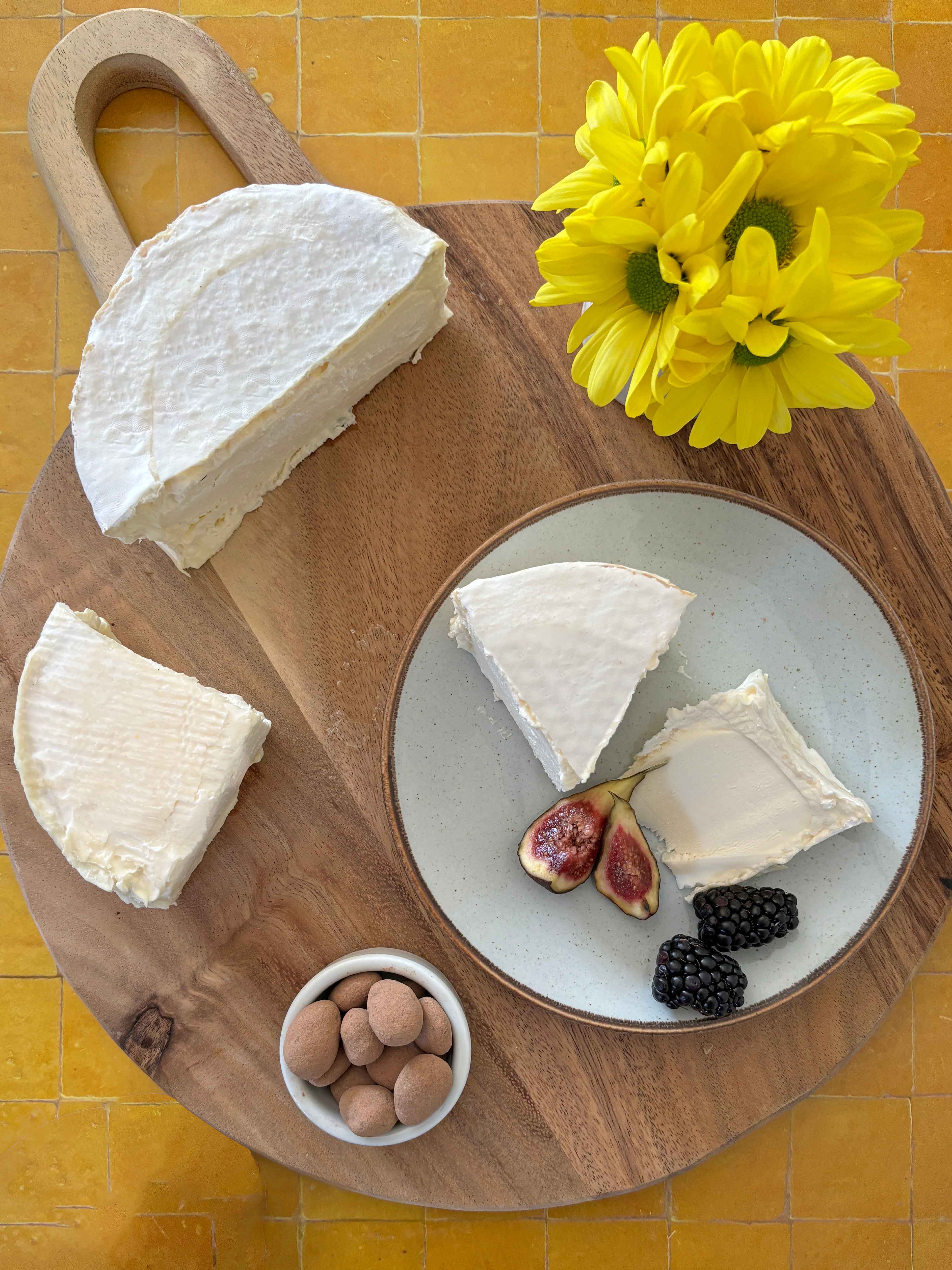 Cheese platter with a wedge of cheese, figs, blackberries, and a small bowl of nuts on a wooden board.