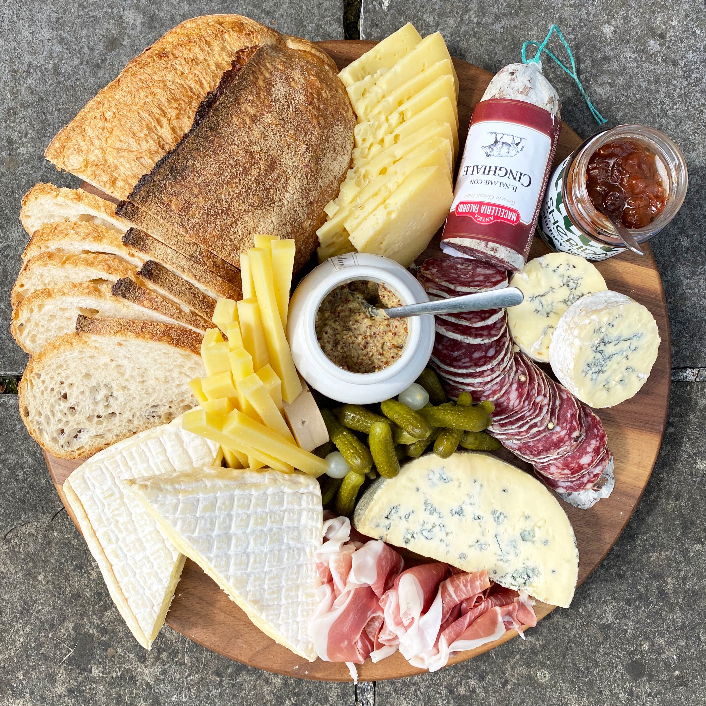 Wooden platter with assorted cheeses, meats, and bread on a concrete surface