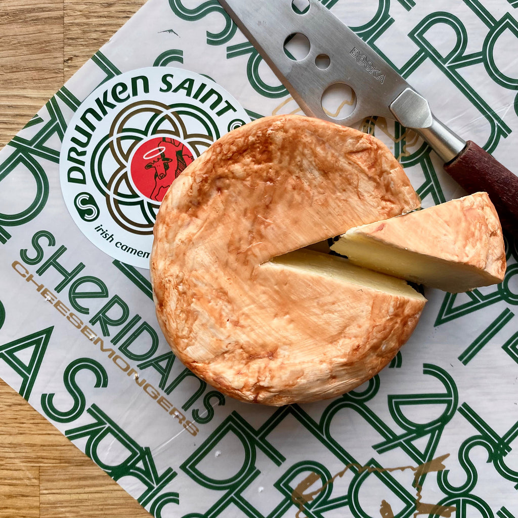 Round of cheese with a slice cut, placed on a Sheridan's Cheeses & Wines branded paper with a knife.