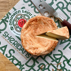 Round of cheese with a slice cut, placed on a Sheridan's Cheeses & Wines branded paper with a knife.