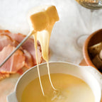 Fork lifting melted cheese from a fondue pot beside sliced ham