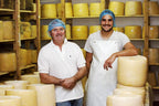 Two men standing in a cheese factory with shelves of cheese behind them.