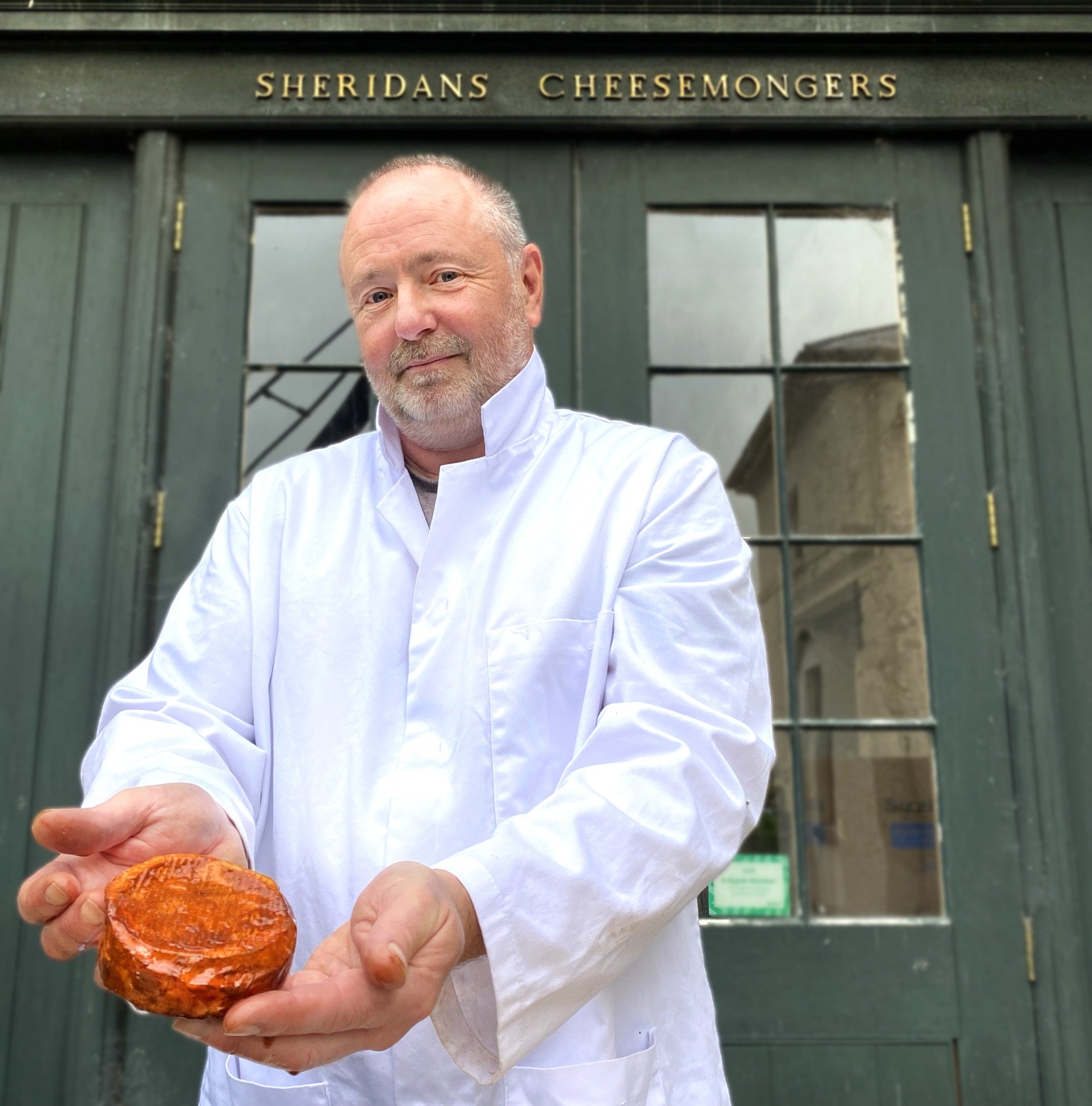 Man in a white coat holding a cheese in front of a building with 'Sheridan's Cheesemongers' sign.