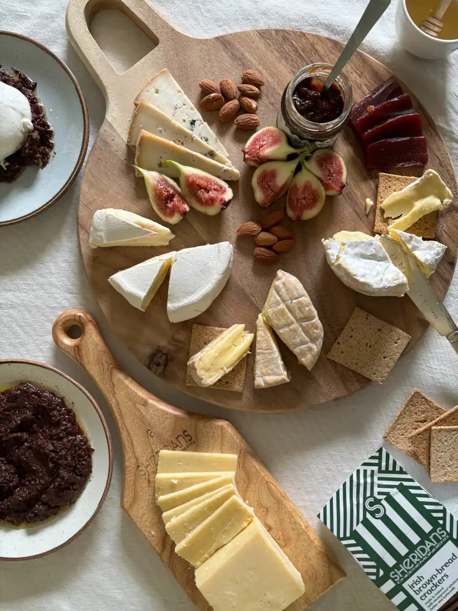 Wooden cheeseboard with various types of cheese, figs, nuts, and crackers on a white tablecloth.