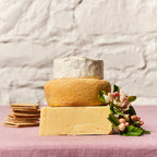 Three types of cheese stacked on a pink surface with crackers and flowers.