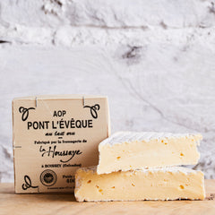 Two pieces of cheese with a cardboard box labeled 'AOP Pont l'Evèque' on a wooden surface.