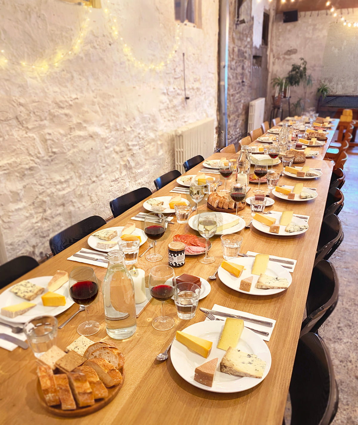 A long wooden dining table set for multiple guests in a rustic indoor venue, with plates of cheese, bread, crackers, wine glasses, and water bottles. White walls, exposed brick, and warm string lights are visible in the background.