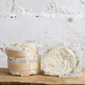 Two wheels of cheese, one in a wooden basket and the other in plastic wrap, on a wooden surface with a light gray textured background.