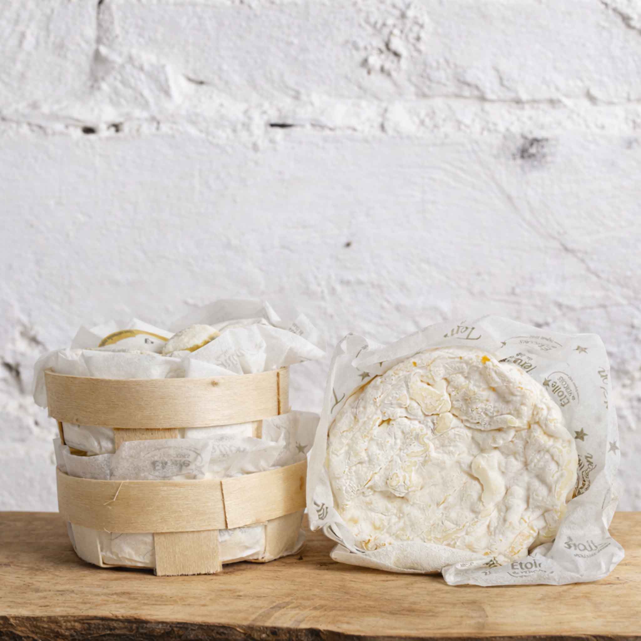 Two wheels of cheese, one in a wooden basket and the other in plastic wrap, on a wooden surface with a light gray textured background.